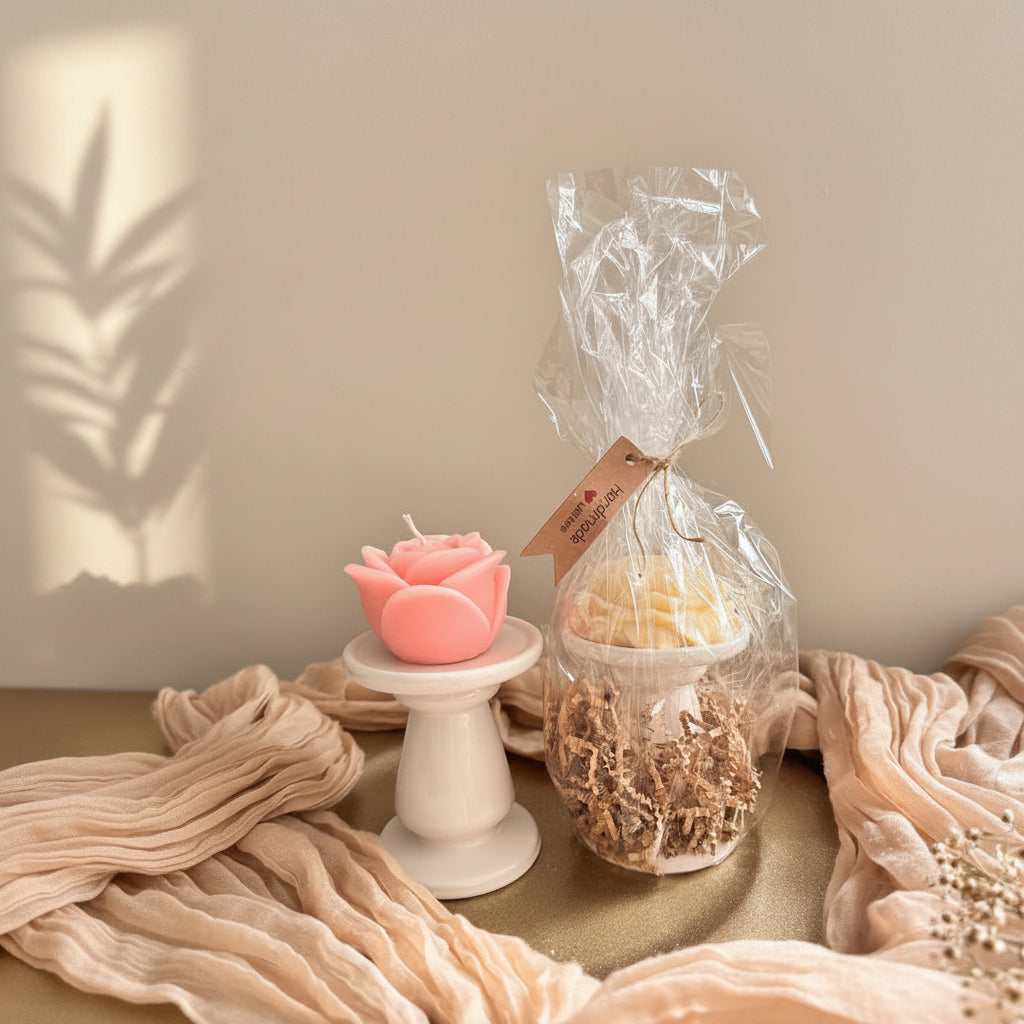 A gift set featuring a pink rose-shaped candle on a small white pedestal and a clear plastic-wrapped jar filled with shredded paper and a yellow item, tied with a tag that reads 'handcrafted with love' and 'Merry & Bright'. The items are styled on a gold surface with beige draped fabric against a gray background.
