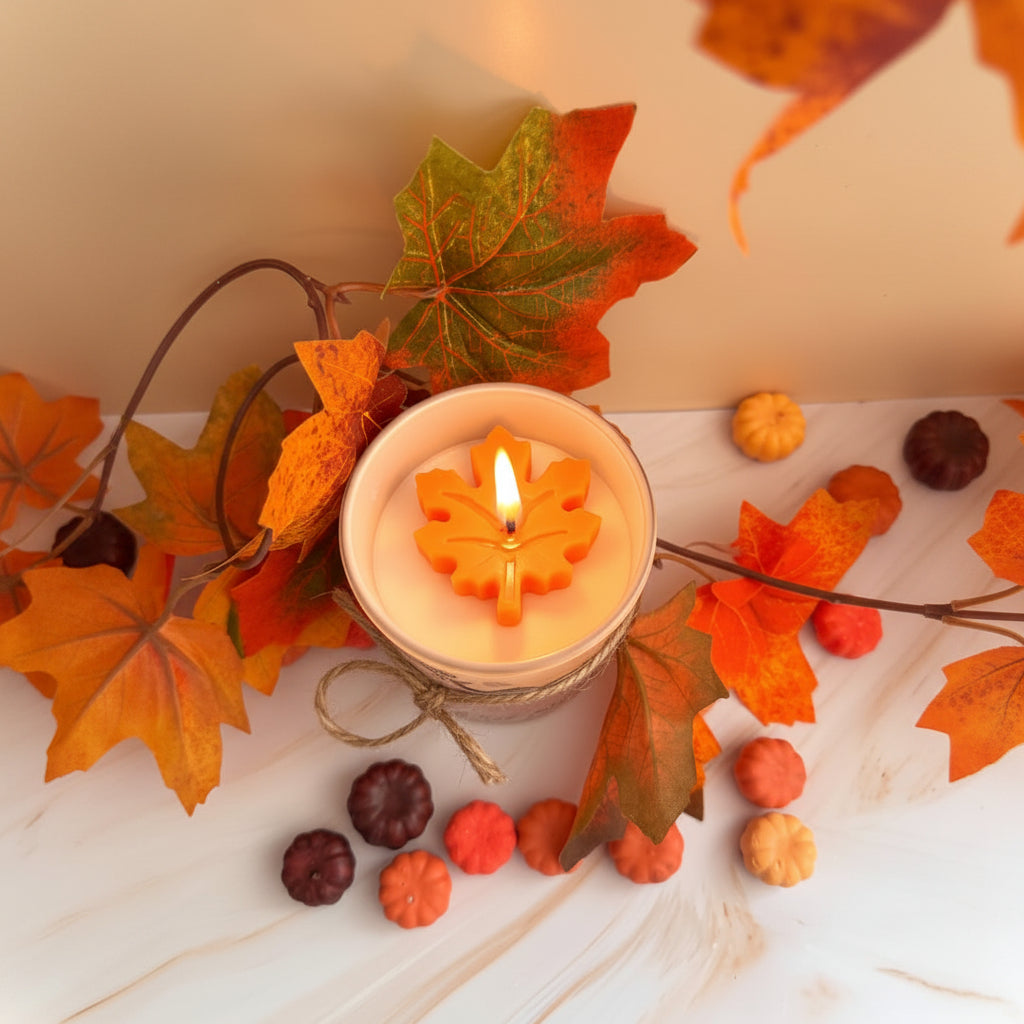 An autumn-themed candle in a clear glass jar with a white wax body and an orange maple leaf-shaped wax float, styled with colorful fall leaves, small pumpkins, and twine on a light marble surface.